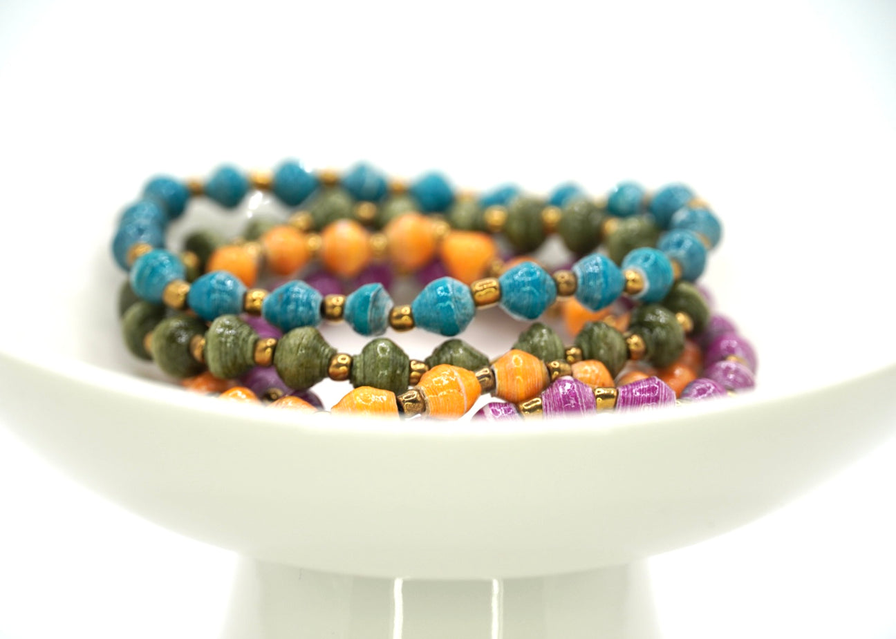 A set of four multicolored paper bead bracelets displayed on a white plate against a white background.